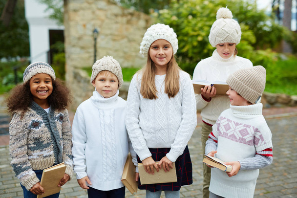 Enfants de différents âges posant à l'extérieur souriant à la caméra et tenant des livres, tous habillés de vêtements en tricot similaires
 - Photo, image