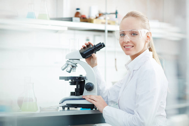 Side view portrait of beautiful blond woman using microscope and looking at camera while working on research in modern laboratory - Photo, Image