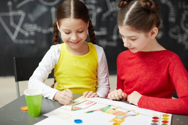 Portrait of two smiling little girls painting pictures together in art studio class sitting against blackboard - 写真・画像