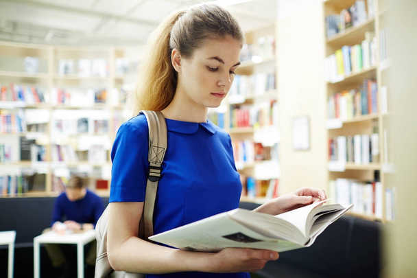 Portret van mooie jonge vrouw lezing boek in bibliotheek permanent door boekenkasten en voorbereiding van de examens - Foto, afbeelding
