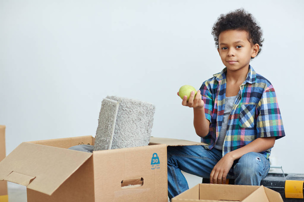 Мальчик с зеленым яблоком сидит за распакованной коробкой
 - Фото, изображение