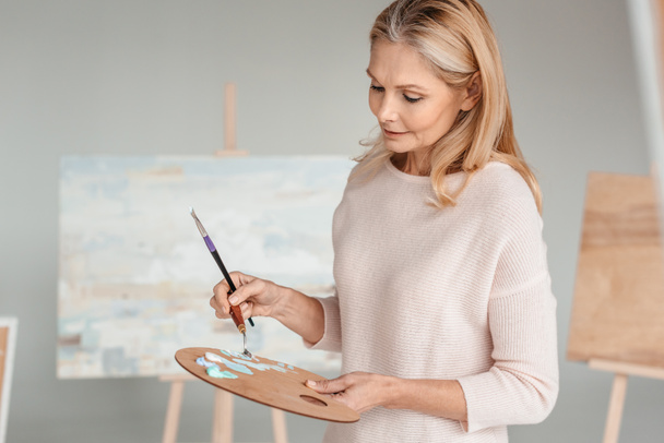 mature woman holding palette and paintbrush in art studio - Фото, изображение