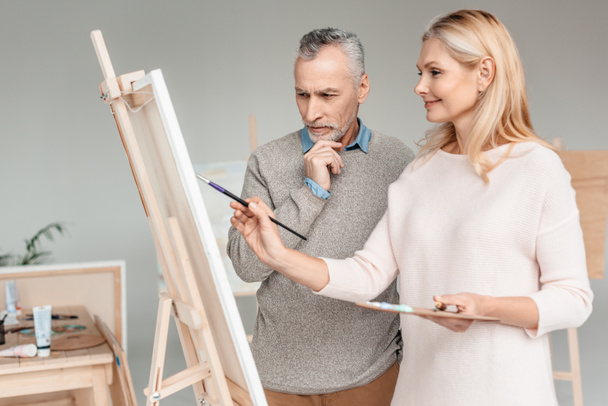 älterer Mann blickt auf Staffelei, während eine ältere Frau lächelt und während des Kunstunterrichts darauf malt - Foto, Bild