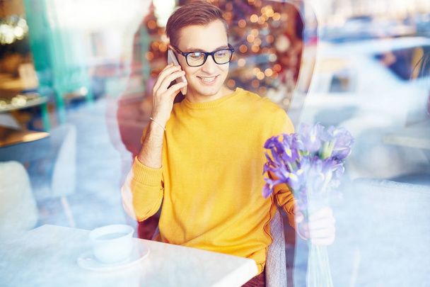 Portrait of smiling young man speaking by phone in cafe while waiting for date with bouquet  of irises - Photo, image
