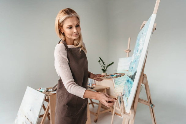 beautiful mature woman in apron painting on easel at art class  - Foto, Bild