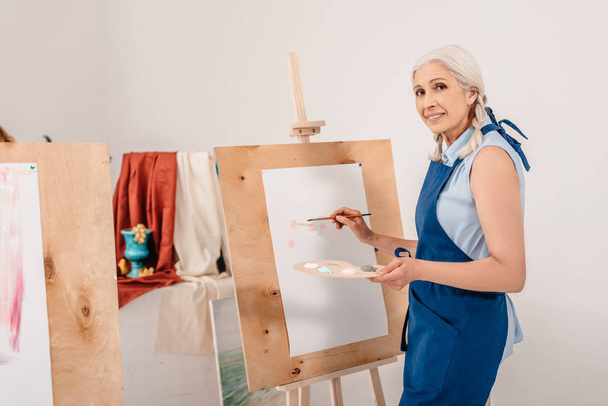 beautiful senior woman smiling at camera while painting on easel in art studio - Fotografie, Obrázek