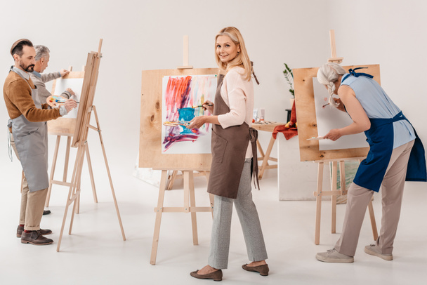 mannelijke en vrouwelijke volwassen studenten in schorten samen schilderen op schildersezels in de kunst klas - Foto, afbeelding