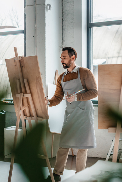 selective focus of mature male artist painting on easel in art studio - Photo, Image