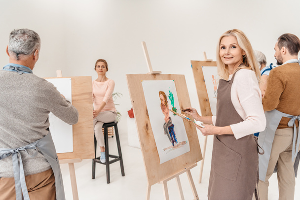 reife Frau lächelt in die Kamera, während sie im Kunstunterricht auf Staffelei malt - Foto, Bild