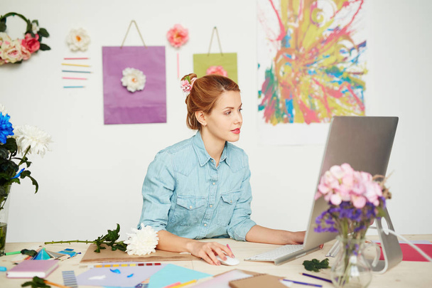 Kaunis graafinen suunnittelija casualwear istuu tietokoneen edessä ja kääritty lupaava projekti, valkoinen seinä koristeltu kukkia ja abstrakti maalaus taustalla
 - Valokuva, kuva