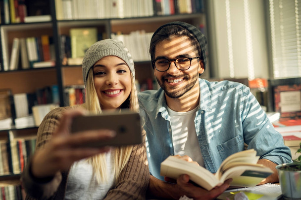 couple of happy young students taking selfie in library - Фото, изображение