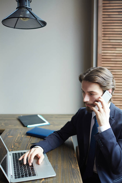 Портрет молодого успішного бізнесмена, який працює з ноутбуком і розмовляє по телефону за дерев'яним столом
 - Фото, зображення