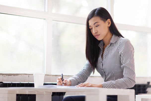 Femme d'affaires asiatique concentrée assise dans un bureau spacieux et prenant des notes avec stylo, portrait taille haute
 - Photo, image