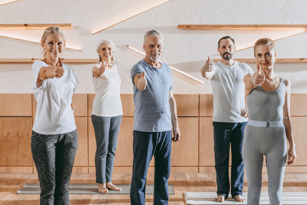 senior people and instructor in sportswear showing thumbs up and smiling at camera in fitness studio - Fotografie, Obrázek