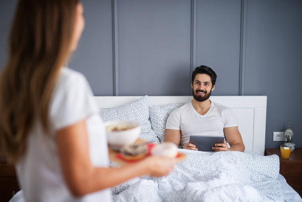 glücklicher bärtiger Mann im Bett mit Tablet-PC, Freundin bringt Frühstück  - Foto, Bild