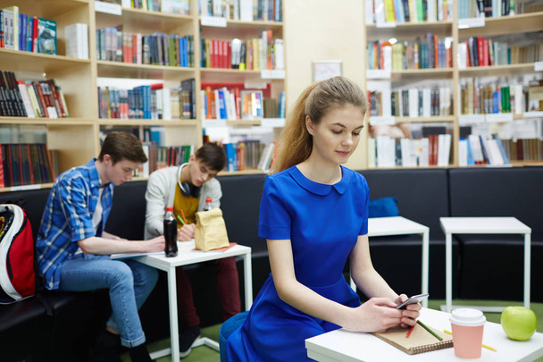Grupo de estudiantes que estudian en la biblioteca sentados en las mesas, se centran en la mujer joven y bonita usando un teléfono inteligente y tomando notas
 - Foto, Imagen