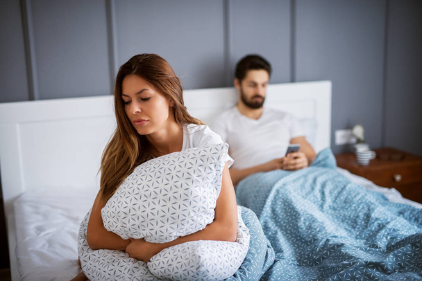 giovane donna triste seduta sul letto e cuscino abbracciato, fidanzato con cellulare in mano sullo sfondo
 - Foto, immagini