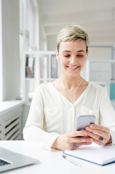 Positieve jonge zakenvrouw kiezen haar favoriete nummers uit de afspeellijst in smartphone door werkplek - Foto, afbeelding