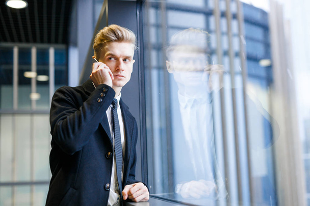 Fiducioso impiegato biondo che telefona al suo cliente mentre sta vicino alla finestra della hall dell'ufficio e distoglie lo sguardo, ritratto in vita
 - Foto, immagini