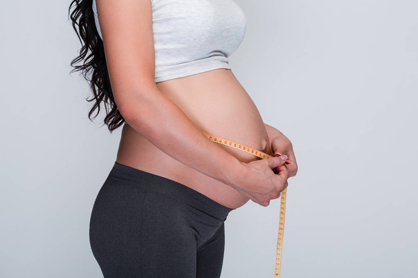 ¡Nuevos centímetros cada mes! Vista lateral parte de una joven embarazada con cinta métrica alrededor de su vientre de pie sobre fondo blanco
 - Foto, imagen