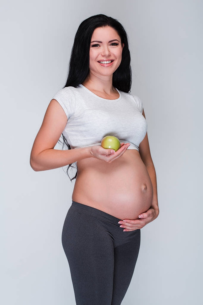 Sağlıklı güzellik. Gülümseme ile kameraya bakarak ve duran yeşil elma beyaz arka plan tutan güzel genç hamile kadın - Fotoğraf, Görsel
