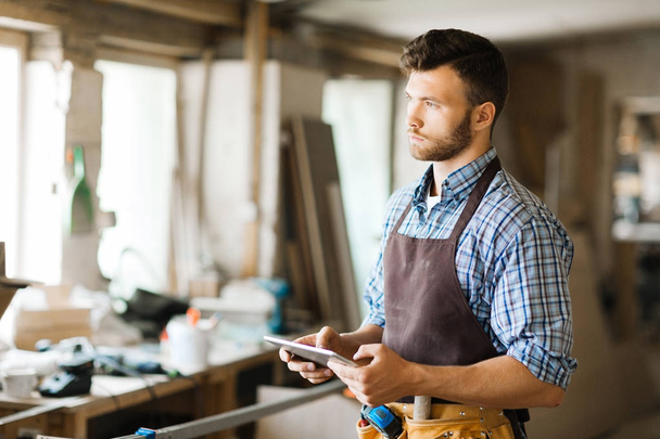 Pensativo joven carpintero sosteniendo tableta digital en las manos mientras mira por la ventana del taller, retrato de cintura hacia arriba
 - Foto, Imagen