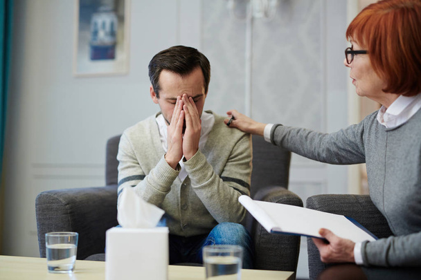 Upset middle-aged patient covering face with hands while sitting on cozy armchair, his mature psychologist trying to comfort him - Photo, Image
