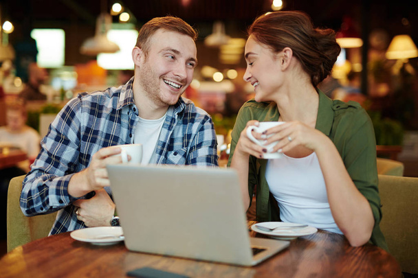 Lovely couple spending evening in cozy small cafe: they enjoying delicious coffee and watching favorite movie on laptop, waist-up portrait - Photo, image