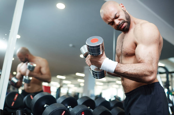 Portrait of shirtless muscular man doing arm exercise working out with dumbbells during strength training in modern gym, flexing and pumping bicep muscles - Photo, Image