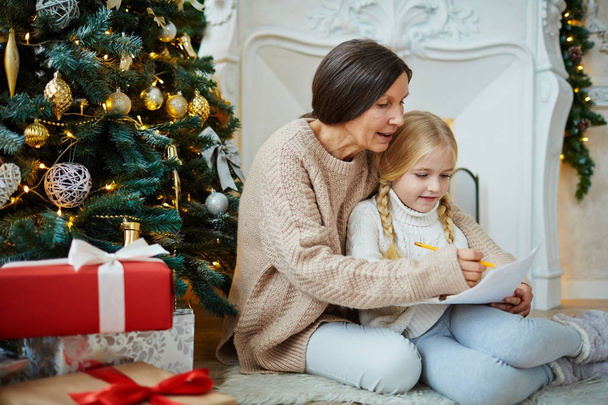 Mature woman and her granddaughter drawing xmas picture by decorated firtree - Foto, Imagem