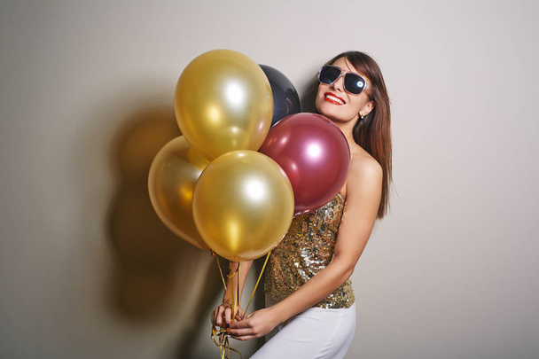 Waist-up portrait of beautiful young woman holding bunch of colorful balloons in hands and looking at camera with toothy smile, studio shot - Photo, Image
