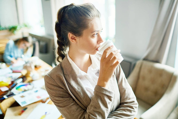 Jovem empresária pensativa bebendo no início do dia de trabalho em seu estúdio
 - Foto, Imagem