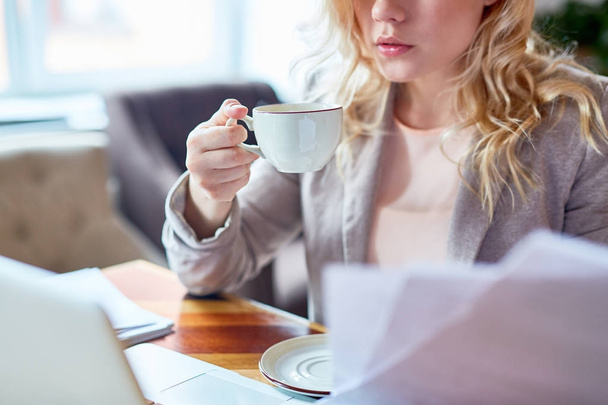 Close-up shot of unrecognizable young entrepreneur enjoying fragrant cappuccino and working on promising project while sitting at cafe table, blurred background - Photo, Image