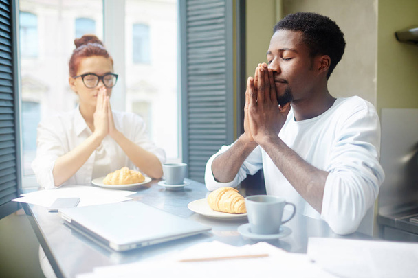 Multi-etnische paar verzameld rond tafel en bidden voordat u ontbijt, huisgemaakte croissants en geurige koffie die hen te wachten - Foto, afbeelding
