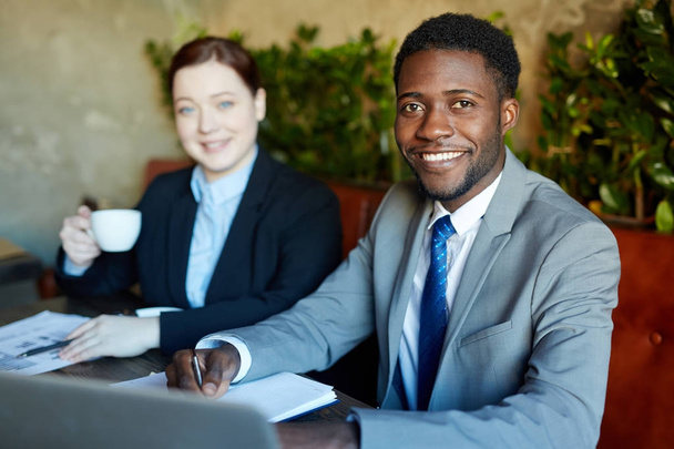Dois empresários durante a reunião de trabalho no café moderno: Bonito empresário afro-americano sorrindo alegremente e olhando para a câmera enquanto trabalhava no laptop com seu colega
 - Foto, Imagem