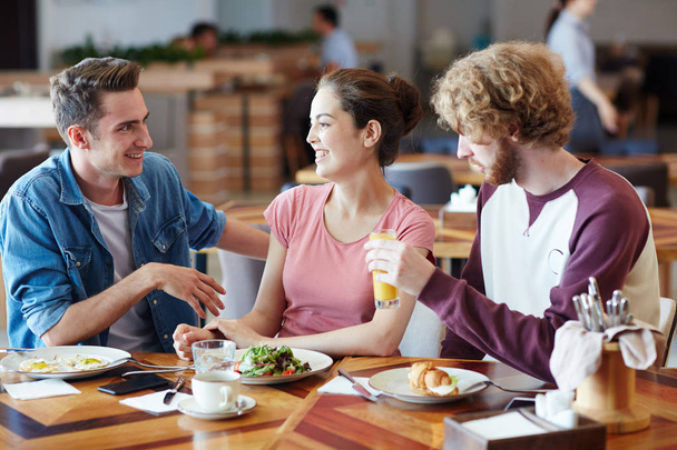 Multi-ethnic group of cheerful friends gathered together at lunchtime in cozy small cafe and chatting animatedly with each other, blurred background - Photo, Image