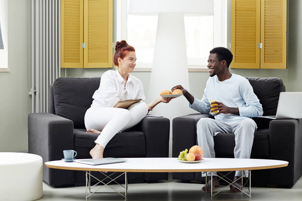 Multi-ethnic young couple enjoying each others company while having breakfast at modern living room, pretty woman passing plate with handmade croissants to her Afro-American boyfriend - Foto, immagini
