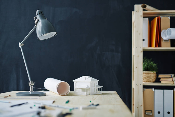 Фон снимок рабочего места дизайнеров с моделью дома на столе, металлической лампой, чертежами и принадлежностями к доске в современном офисе, копировальное пространство
 - Фото, изображение