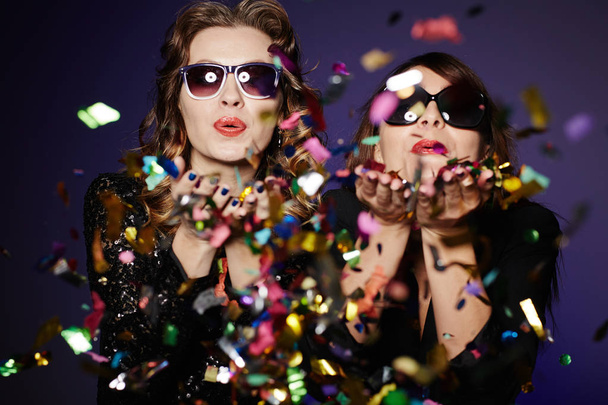 Plezier met beste vriend: twee stijlvolle vrouwen permanent tegen de donkere achtergrond en kleurrijke confetti, taille-omhoog blazen groep portret - Foto, afbeelding