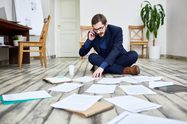 Portrait of middle aged bearded man speaking by phone and sorting documents sitting on floor in office - 写真・画像