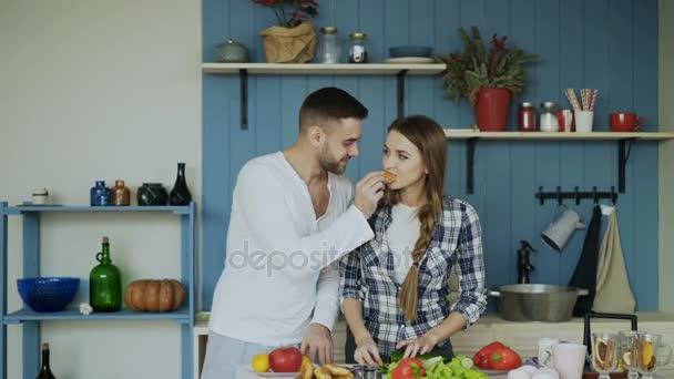 Happy young couple in the kitchen. Attractive dancing man feeding his girlfriend while she is cooking in the morning - Кадри, відео