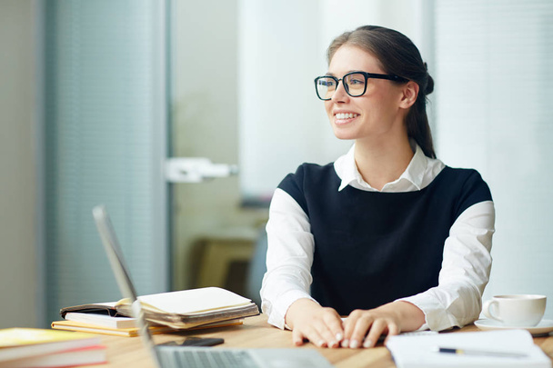 Waist-up portrait of attractive young manager with charming smile looking away while sitting at office desk, blurred background - Φωτογραφία, εικόνα
