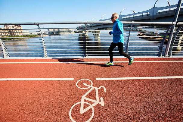 Профиль пожилого спринтера, бегущего вдоль атлетического трека во время подготовки к марафону, безоблачного голубого неба и глубокого озера на заднем плане
 - Фото, изображение
