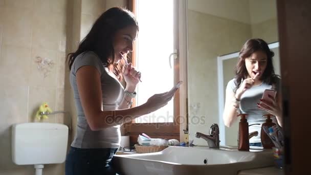 Молодая красивая женщина моет зубы, разговаривая смартфон в помещении в ванной комнате - коммуникация, технологии, концепция зубной щетки
 - Кадры, видео