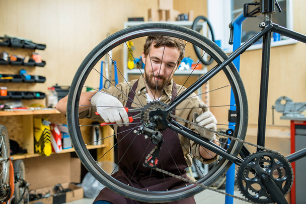 Энтузиастичный молодой механик в фартуке сидит на ягодицах и регулирует велосипедную цепь, интерьер современной мастерской на заднем плане
 - Фото, изображение