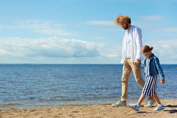 Молодой человек и его дочь держатся за руки, прогуливаясь по берегу моря в солнечный день
 - Фото, изображение