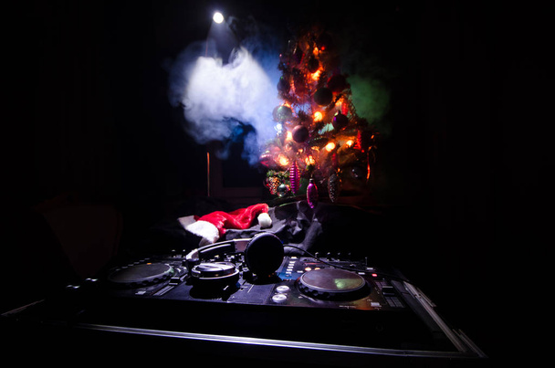 Dj mixer with headphones on dark nightclub background with Christmas tree New Year Eve. Close up view of New Year elements or symbols (Santa Clause, Snowman, Dog 2018, gift box) on a Dj table. toned - Fotó, kép