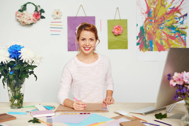 freudige Modedesignerin mit breitem Lächeln, die für Fotos posiert, während sie den Bleistift in der Hand hält, um im Notizbuch zu skizzieren - Foto, Bild