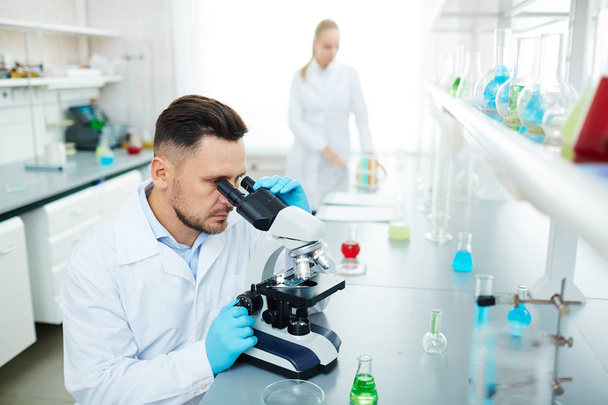 Porträt von Wissenschaftlern bei der Arbeit im modernen Labor: Mann mikroskopiert am Tisch mit Frau reaktfreie Flüssigkeit im Reagenzglas - Foto, Bild