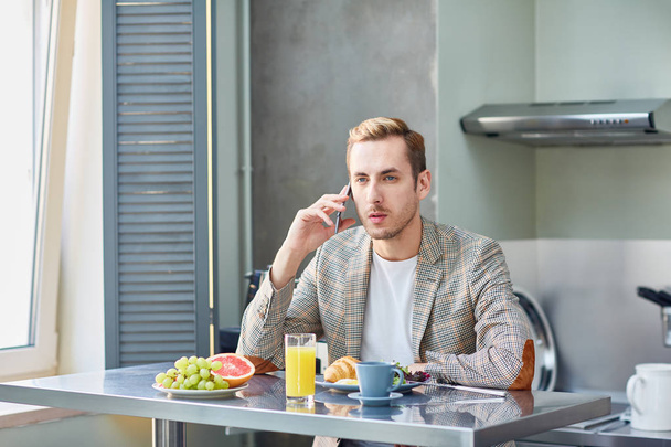 A fiatalember szolgált asztalnál ült, és beszél a smartphone a konyhában - Fotó, kép